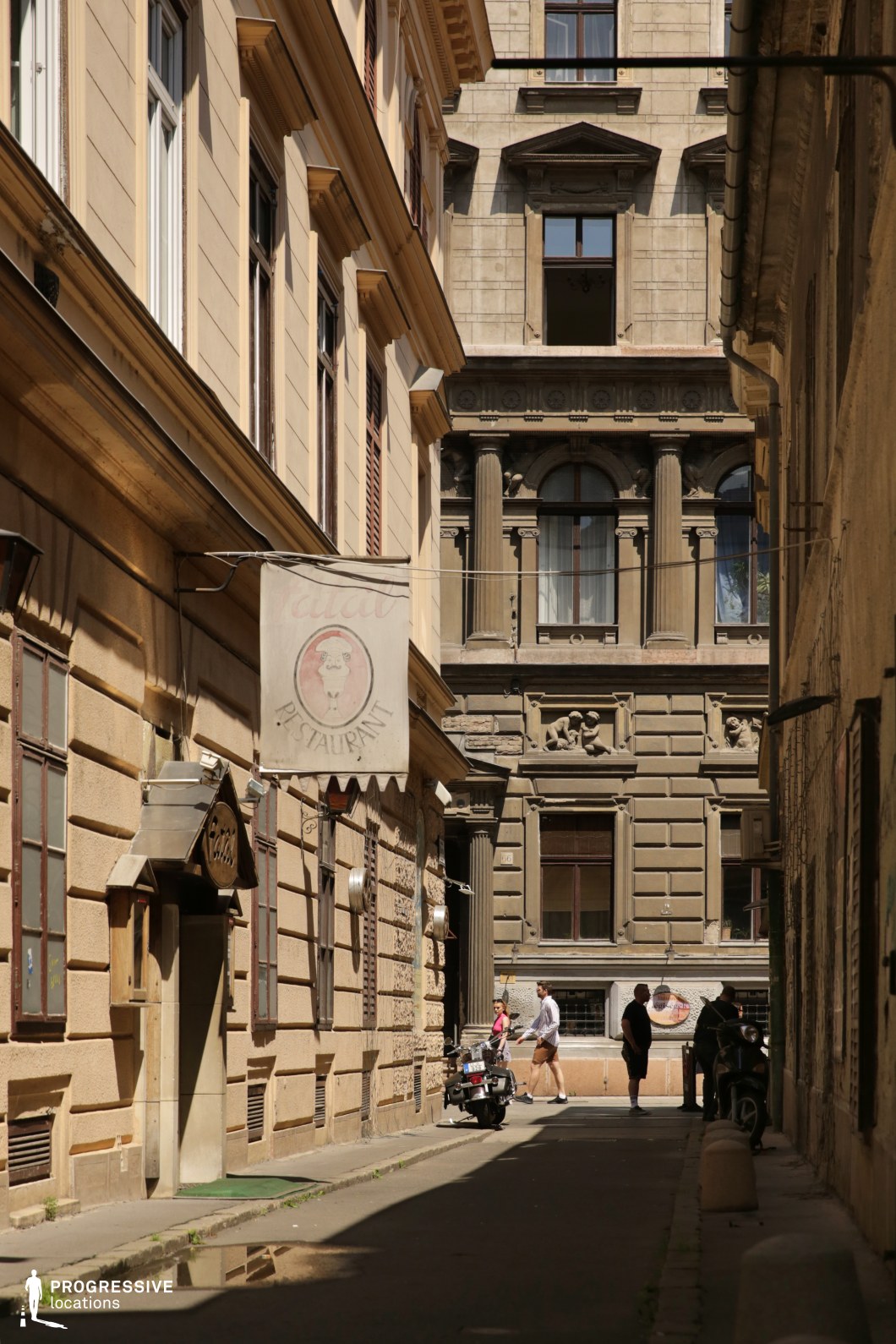 This is a narrow, intimate atmosphere of a historic urban alley, framed by 19th-century classical architecture with warm beige tones and detailed stone façades. 