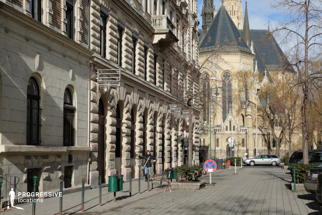 The image captures a visually striking urban scene featuring a blend of historic and functional design elements. The dominant color palette includes light brown and gray tones on the building façades and paved ground, contrasted by the darker slate-blue roofing of the neo-Gothic church in the background. These hues create a harmonious yet distinguished atmosphere that exudes historical richness. The materials include finely carved stone on the arched façade of the building, decorative masonry, and detailed wrought ironwork on the windows. The church’s towering spires and pointed arches, adorned with intricate stone carvings, emphasize its neo-Gothic architectural character, adding depth and elegance to the composition. The street is relatively narrow, lined with small planters, benches, and low barriers, which contribute to a cozy and semi-enclosed ambiance. The perspective naturally leads the eye along the sidewalk, with the prominent church standing as a focal point in the background. Functional urban elements, such as trash bins, benches, and signage, provide a modern and practical layer to the historic setting. The combination of architectural sophistication and usability makes this location suitable for creative projects requiring a balance of old-world charm and urban convenience.