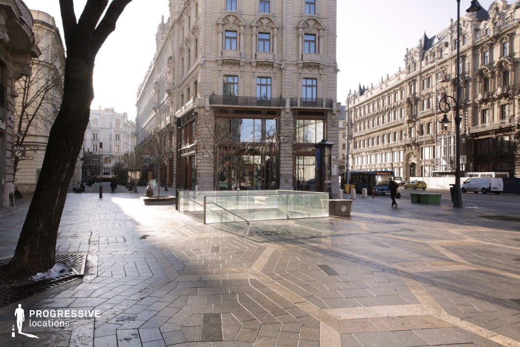 The square exudes a refined cosmopolitan ambiance, blending Parisian elegance with a hint of New York sophistication. Encircled by elegant buildings, the space is characterized by a harmonious color palette of soft beige, cream, and light grey, accented by intricate decorative stonework, wrought-iron balconies, and expansive glass windows that embody the sophistication of classical European architecture. At the heart of the square lies a glass structure—a sleek, modern entrance to an underground passage or metro access. This functional feature not only serves as a practical element of urban infrastructure but also opens the setting to scenes related to the bustling, layered dynamics of city life. The glass design adds a contemporary urban contrast to the historical backdrop, making it a striking visual centerpiece that bridges tradition and modernity. The geometric tiling of the paved square, with its muted greys and earthy tones, complements the historic façades and creates a seamless, polished appearance. The black cast-iron street lamps add an additional layer of elegance, their intricate craftsmanship standing out against the neutral tones of the architecture. These lamps, along with the minimalist landscaping, underscore the sophisticated design ethos of the area. This location is particularly suited for productions seeking to capture the intersection of urban functionality and timeless charm. The underground entrance introduces opportunities for scenes depicting the bustling rhythms of city life—be it a chance encounter, a hurried commute, or a dramatic pursuit. The color scheme and urban composition make it an ideal setting for commercials, fashion editorials, or cinematic productions aiming to merge historic beauty with modern vitality.