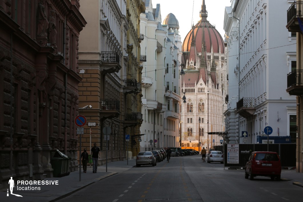 The image depicts a narrow urban street framed by ornate classical-style buildings, leading toward a view of the iconic dome and spires of a grand parliament building in the background. The architectural details of the street showcase a mix of rich, textured façades on the left with dark red tones, and lighter, more refined façades on the right, blending historic and elegant urban design. The street is lined with small balconies adorned with wrought iron railings, adding a sense of charm and European sophistication. The perspective emphasizes the gradual transition from the shadowed, intimate atmosphere of the street to the brightly lit, majestic architecture of the parliament building. The dome, with its intricate Gothic detailing and reddish-brown color, dominates the background and serves as the focal point of the composition. The pavement includes a subtle bike lane, creating a sense of functionality within the historical setting. The overall view combines the narrow, enclosed feeling of the street with the openness and grandeur of the distant landmark, making this location ideal for cinematic scenes that require a balance of intimate urban character and impressive architectural grandeur. This scene is best suited for wide-angle shots that highlight the contrast between the close, richly detailed buildings and the monumental parliament structure in the background.