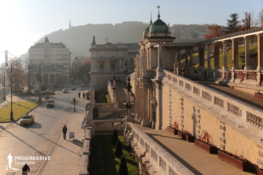 This is an elegantly designed terrace overlooking a historic urban street, framed by ornate architectural details and a gently sloping hillside in the background. 