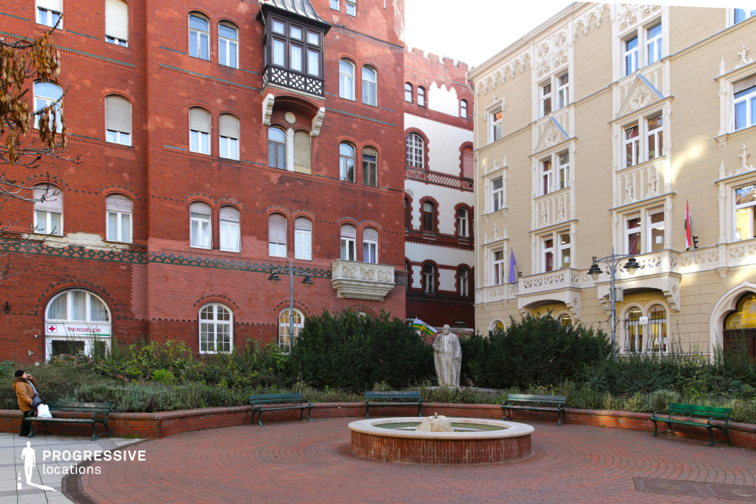 This image showcases a charming urban square framed by diverse architectural styles, creating a lively and visually engaging atmosphere.

At the center of the square is a circular fountain with a brick base, surrounded by curved benches and meticulously arranged greenery. A small statue within the landscaped area adds cultural and artistic significance to the setting. The layout encourages relaxation and interaction, making the space feel both welcoming and functional.

The dominant materials include red brick, prominently featured in the façade of the taller building on the left and the paving of the square itself. Adjacent to it, a beige-colored building with ornate stucco details and decorative balconies contrasts with the more industrial character of the red brick. The window designs range from rounded arches on the brick structure to rectangular frames with elaborate trims on the beige building, blending modernity and tradition.

The spatial arrangement is compact yet open, with the circular design of the fountain and benches drawing attention to the square's central features. The greenery, brick paving, and sculptural elements enhance the square’s aesthetic while emphasizing its multifunctional urban role.

This square offers an inviting and versatile location, perfect for productions that require a balance of historic charm and urban vibrancy. Its architectural diversity and carefully designed public space make it ideal for scenes emphasizing culture, relaxation, or city life.