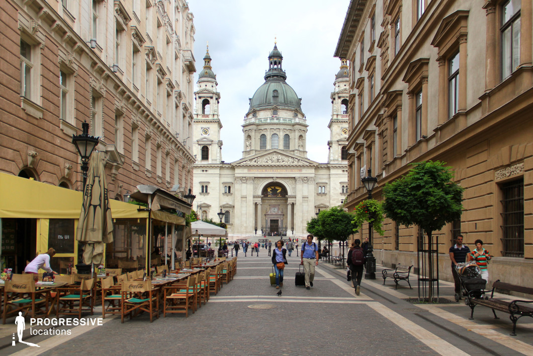 This image captures a lively pedestrian street framed by classical architecture, culminating in the view of a grand basilica with twin towers and a central dome. The atmosphere is urban yet inviting, with a mix of activity and historical grandeur.

The color palette prominently features beige and light brown tones from the surrounding buildings, complemented by the dark green dome and the white stone façade of the basilica. The yellow awnings of the café add a splash of vibrancy to the scene, while the cobblestone paving provides a textured and authentic touch.

The materials include finely crafted stone and stucco on the buildings, detailed wrought iron for streetlamps, and wooden furniture for the outdoor dining area. The basilica stands as the focal point with its neoclassical design, featuring intricate carvings, columns, and arched windows, exuding timeless elegance.

The spatial arrangement emphasizes the long, straight perspective of the pedestrian street, leading the viewer’s eye directly to the basilica. The arrangement of café seating and small ornamental trees on either side adds symmetry and a sense of community.

This location offers a picturesque blend of historic and urban elements, ideal for productions or photographs that highlight cultural richness, architectural sophistication, and a bustling yet organized city environment.