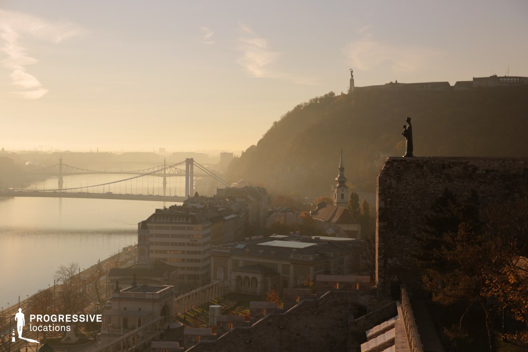 This image presents a breathtaking panorama of a riverside city, dominated by the tranquil water of the river, a historic hill with a prominent statue, and a bridge stretching elegantly into the distance. The golden tones of the light envelop the scene, adding a soft and atmospheric quality. The color palette is composed of muted gold and warm earthy tones, blending seamlessly with the natural greens of the hillside and the subtle gray-blue of the river. The light haze over the scene enhances its depth, creating a dreamy, timeless ambiance. The materials in the foreground include rough stone from the historic walls and structures, contrasted with the sleek design of the bridge in the background. The hill features a prominent statue standing atop a dramatic outcrop, while a spire of a church adds vertical balance to the composition, symbolizing the city's cultural and historical significance. The perspective offers a commanding view from an elevated vantage point, capturing the layered complexity of the city, the flowing river, and the connection provided by the bridge. The composition naturally guides the viewer's gaze from the foreground details to the distant urban skyline and the hazy horizon. This location is ideal for productions or photography that aim to highlight the serene beauty of urban and natural landscapes, with a strong emphasis on cultural landmarks and a contemplative atmosphere. The combination of historic and modern elements makes it suitable for a wide range of visual narratives.