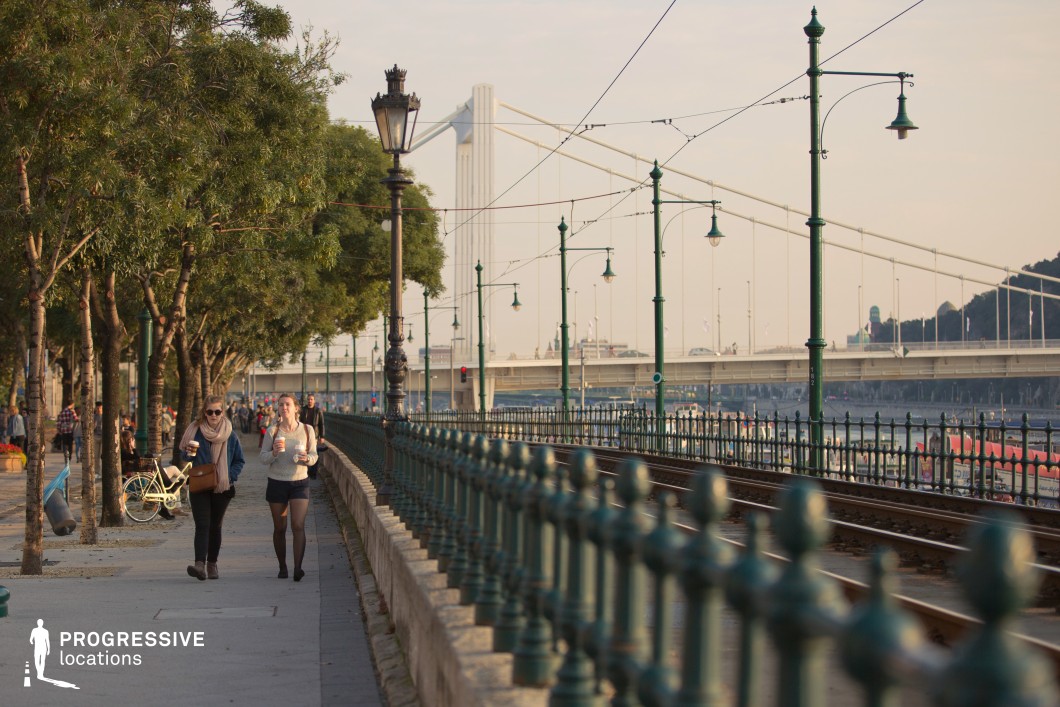 This image captures a scenic riverside promenade lined with trees, iron railings, and a tram line, leading to a striking view of a modern suspension bridge in the background. The composition combines natural and urban elements, creating a dynamic and inviting atmosphere. The dominant colors include green tones from the railings and trees, beige hues from the pavement, and the soft gray of the bridge structure. The warm lighting highlights the textures and details, adding a golden hue to the scene. The materials featured include wrought iron railings with decorative elements, asphalt and stone for the walkway and tram tracks, and steel for the suspension bridge. The row of streetlamps adds a vintage charm to the modern riverside setting. The perspective emphasizes the linear elements of the tram tracks and promenade, naturally guiding the viewer's eye toward the bridge and horizon. The parallel alignment of the tracks, railings, and trees creates a balanced and harmonious composition. This location is ideal for productions or photography highlighting urban life along a riverfront. The combination of historic design elements, modern infrastructure, and lush greenery makes it suitable for scenes requiring a blend of activity, leisure, and scenic beauty.