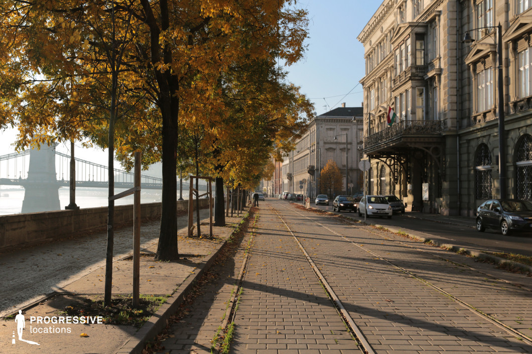 This image portrays a serene riverside promenade flanked by an elegant row of historic buildings on one side and a view of the river on the other. The architectural style of the buildings is classically European, featuring ornate balconies, stone facades, and tall windows. A metal canopy adds further detail to the building in the foreground. The promenade itself includes a tree-lined walkway, cobblestone paths, and tram tracks embedded in the pavement, creating a sense of depth and perspective.

On the left, the view opens to a bridge with a prominent, classical design, featuring large stone pillars and metal cables stretching across the river. The trees along the path, with their foliage providing a natural canopy, add warmth and vibrancy to the urban landscape. This setting seamlessly combines urban elegance with riverside tranquility, offering a perfect backdrop for cinematic scenes or architectural studies.