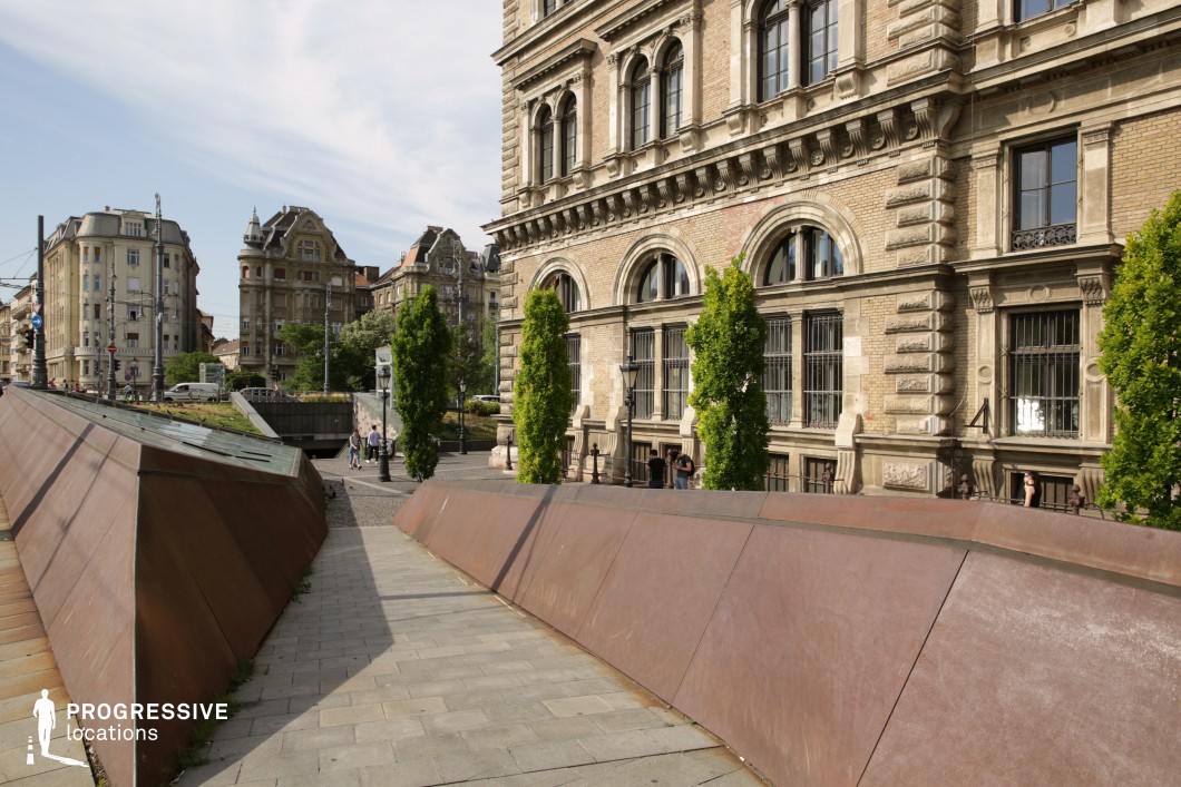 This image presents a visually striking urban setting where modern and historic architectural elements coexist harmoniously. The foreground features a large, rust-colored steel structure with angular, sloped surfaces, creating a narrow passageway that adds a sense of dynamism and directionality. This structure not only contrasts with the Neo-Renaissance building in the background but also provides a perfect setting for action-oriented sequences such as skateboarding, rollerblading, or cycling, thanks to its sleek design and spacious layout. In the background, a small underpass or tunnel adds an additional layer of depth and visual interest, hinting at connectivity to other parts of the urban environment. The combination of the bold, modern steel elements, the open pedestrian paths, and the stately historic architecture creates a versatile location. The earthy tones of the steel, the light beige bricks, and the green trees provide a balanced and engaging color palette, making this space ideal for productions requiring dynamic urban motion or a mix of old and new architectural styles.