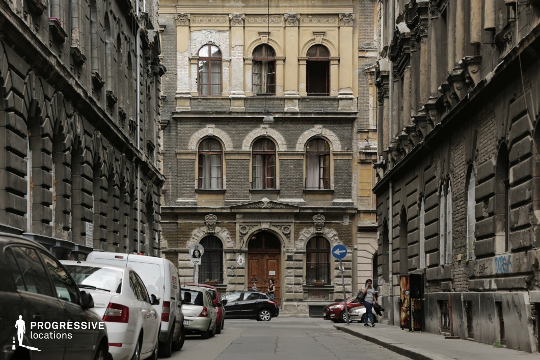This image captures a narrow urban street flanked by tall, ornate tenement buildings. The architecture is a mix of yellow and grey facades with arched windows and decorative stonework, showcasing a late 19th-century or early 20th-century design. The central building features intricate carvings above its main entrance, which is framed by columns and topped with classical motifs. The alley creates a focused, enclosed perspective, emphasizing the verticality of the buildings and the urban density. The aged textures and weathered surfaces of the facades evoke a historical atmosphere, making this location suitable for urban period storytelling or architectural exploration.