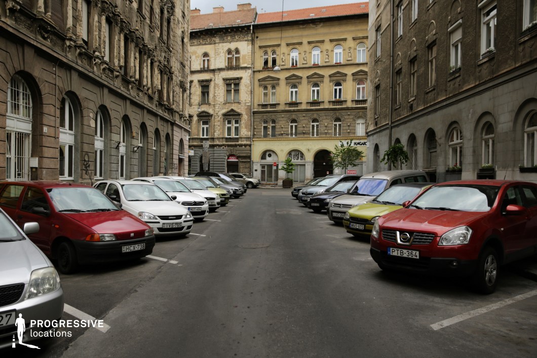 This image depicts an urban street flanked by aging tenement buildings, their facades showcasing a mix of classical details and visible wear. The yellow and beige tones of the central building, with its symmetrical windows and subtle ornamentation, stand out amidst the darker and weathered facades of the neighboring structures. The alignment of the parked vehicles along the street emphasizes its residential nature and enclosed layout. The overall scene reflects a combination of urban density and historical character, evoking a gritty yet nostalgic cityscape that would be ideal for capturing a sense of realism or vintage urban charm.