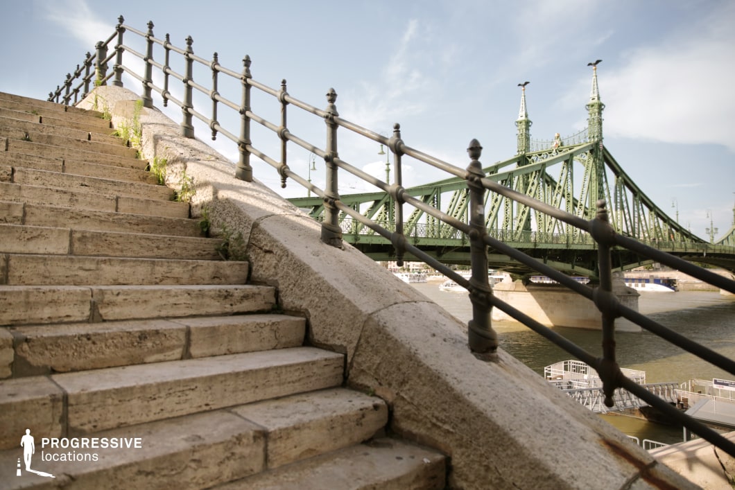 This image features a stone staircase leading upward, framed by a wrought-iron railing, with the iconic green iron bridge as a striking backdrop. The weathered texture of the stone steps, combined with the small patches of greenery sprouting between the cracks, gives the scene a sense of historical character and rugged charm. The low-angle perspective emphasizes the staircase and railing, creating a sense of depth while opening up the frame to the expansive blue sky above. This composition provides a dramatic and visually dynamic setting, often favored in fashion photography, particularly for showcasing sport or outdoor wear, as the upward angle and open space convey a sense of energy, movement, and freedom. The combination of aged materials, industrial elements, and a spacious backdrop makes this location ideal for productions looking for a versatile urban setting with both historical and modern appeal.