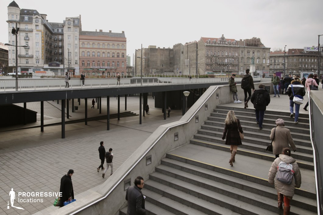 This modern urban space is defined by clean lines and a minimalist architectural approach, dominated by neutral greys and muted beiges. The smooth concrete surfaces of the stairs and ramps are complemented by dark metal railings and columns, creating a contemporary, utilitarian aesthetic. The adjacent historic buildings in the background feature late 19th to early 20th-century eclectic and neoclassical facades, characterized by intricate detailing, decorative cornices, and large windows. The juxtaposition of styles evokes a transitional urban atmosphere, reminiscent of central European cityscapes such as Vienna or Berlin. The subdued color palette emphasizes functionality while providing a subtle contrast to the historic surroundings.