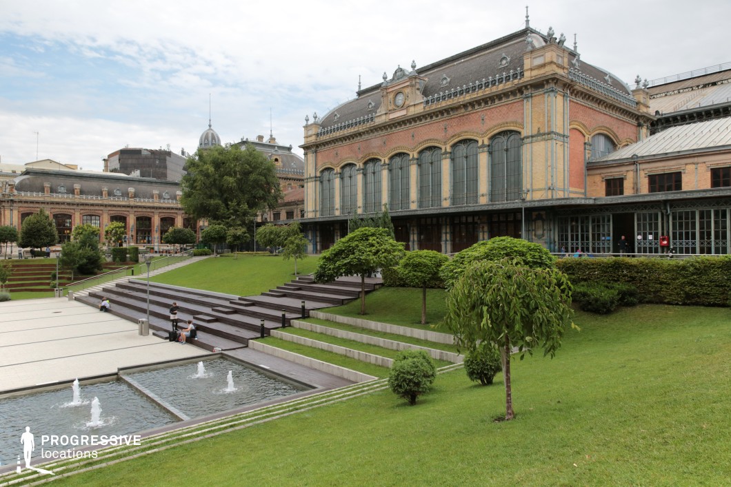 This image showcases an elegant historic building with a striking combination of red brick and pale stone accents, framed by tall arched windows and decorative ironwork. The structure, reminiscent of a classic European railway station or cultural hall, features ornate architectural details such as cornices, balustrades, and a central clock. The building is complemented by a modern, landscaped park in the foreground, characterized by lush green lawns, tiered terraces, and neatly trimmed trees.

The park incorporates clean, geometric design elements, including a series of fountains with rectangular basins, adding a dynamic visual contrast to the historic architecture. The terraced layout provides depth to the scene, creating natural seating areas and pathways that guide the viewer’s gaze toward the building. This location blends the charm of a historic landmark with the functionality and aesthetic of contemporary urban landscaping, making it ideal for versatile outdoor scenes or architectural-focused productions.