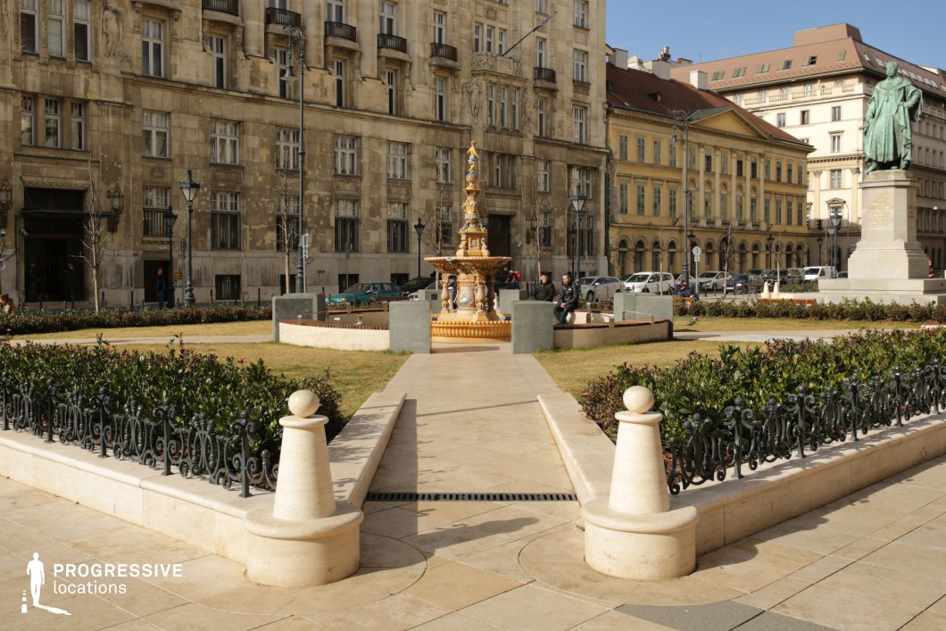 The image captures an elegant urban park surrounded by stately historic buildings. At the center of the park is an ornate fountain, featuring intricate carvings and a golden hue that makes it a standout element of the composition. To the right, a bronze statue of a historical figure on a pedestal adds to the park’s monumental and refined atmosphere.

The park’s layout is symmetrical and meticulously designed, with neatly trimmed hedges and small grassy areas bordered by low, decorative wrought-iron fencing. Paved walkways made of light-toned stone lead visitors through the park, creating a balanced and harmonious structure. The use of light stone in the pavement and fencing posts complements the neutral tones of the surrounding architecture.

The buildings enclosing the park are constructed in a classic European style, with detailed façades, arched windows, and modest balconies, evoking a sense of timelessness and sophistication. The combination of the park's decorative elements and the stately backdrop makes this location ideal for photography or filming scenes requiring an elegant, urban European setting.

The openness of the space, coupled with its rich architectural details, allows for wide-angle shots that emphasize the symmetry of the design, while the ornate fountain and statue serve as focal points for creative visual compositions.