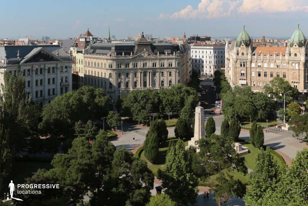 This elegant aerial view captures Szabadság Square, framed by grand historic buildings and lush greenery in a sophisticated urban setting. The surrounding buildings, primarily five to six stories tall, create a consistent sense of scale and emphasize the square's metropolitan character. On the left, a neoclassical-style structure with ornate detailing and a gabled roof adds a touch of refinement, while on the right, a richly decorated building featuring distinctive green domes enhances the area’s grandeur. The square’s centerpiece is a tall obelisk monument, surrounded by symmetrical pathways, manicured hedges, and mature trees, providing a balance of urban sophistication and natural tranquility. This location is ideal for showcasing the grandeur and vibrancy of a historic city center.