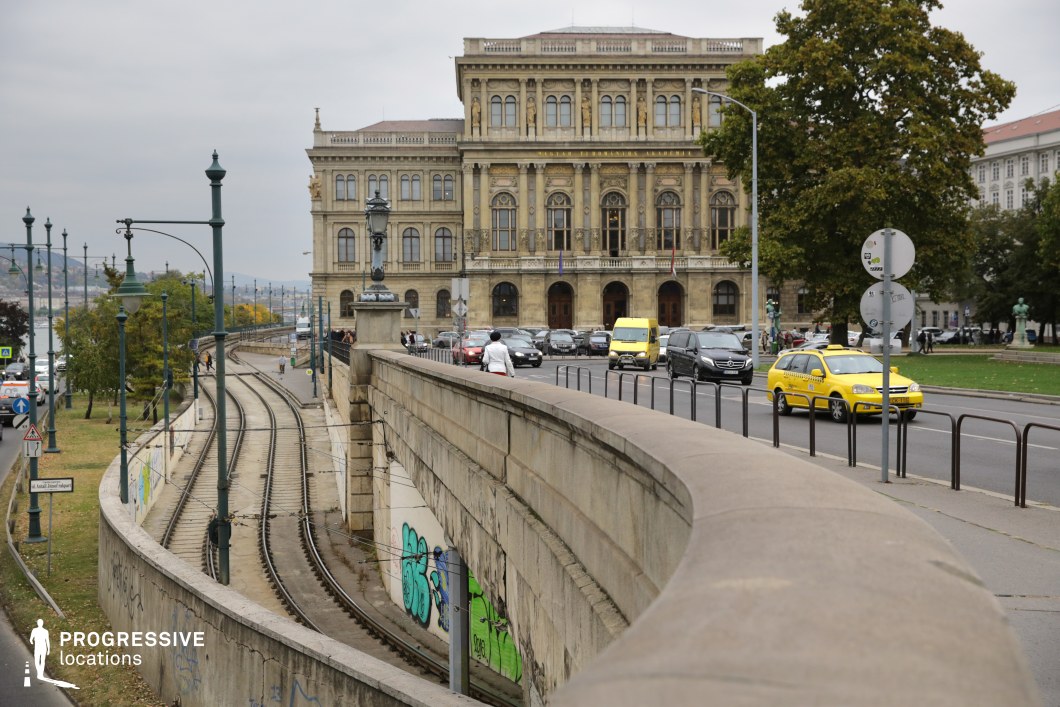This scene blends historic and modern urban elements, with a tram track curving beside a neoclassical building. The building, constructed in the mid-to-late 19th century, features a symmetrical facade, grand arched windows, and Corinthian pilasters, showcasing the elegance of academic classicism. The curved underpass wall introduces a contrasting industrial tone, with graffiti adding a gritty, modern edge. The earthy stone hues and soft greys of the infrastructure form a muted color scheme, which harmonizes with the greenery and distant views of the river. This setting recalls the dynamic contrasts seen in cities like Budapest or Vienna, balancing historic prestige with urban functionality.