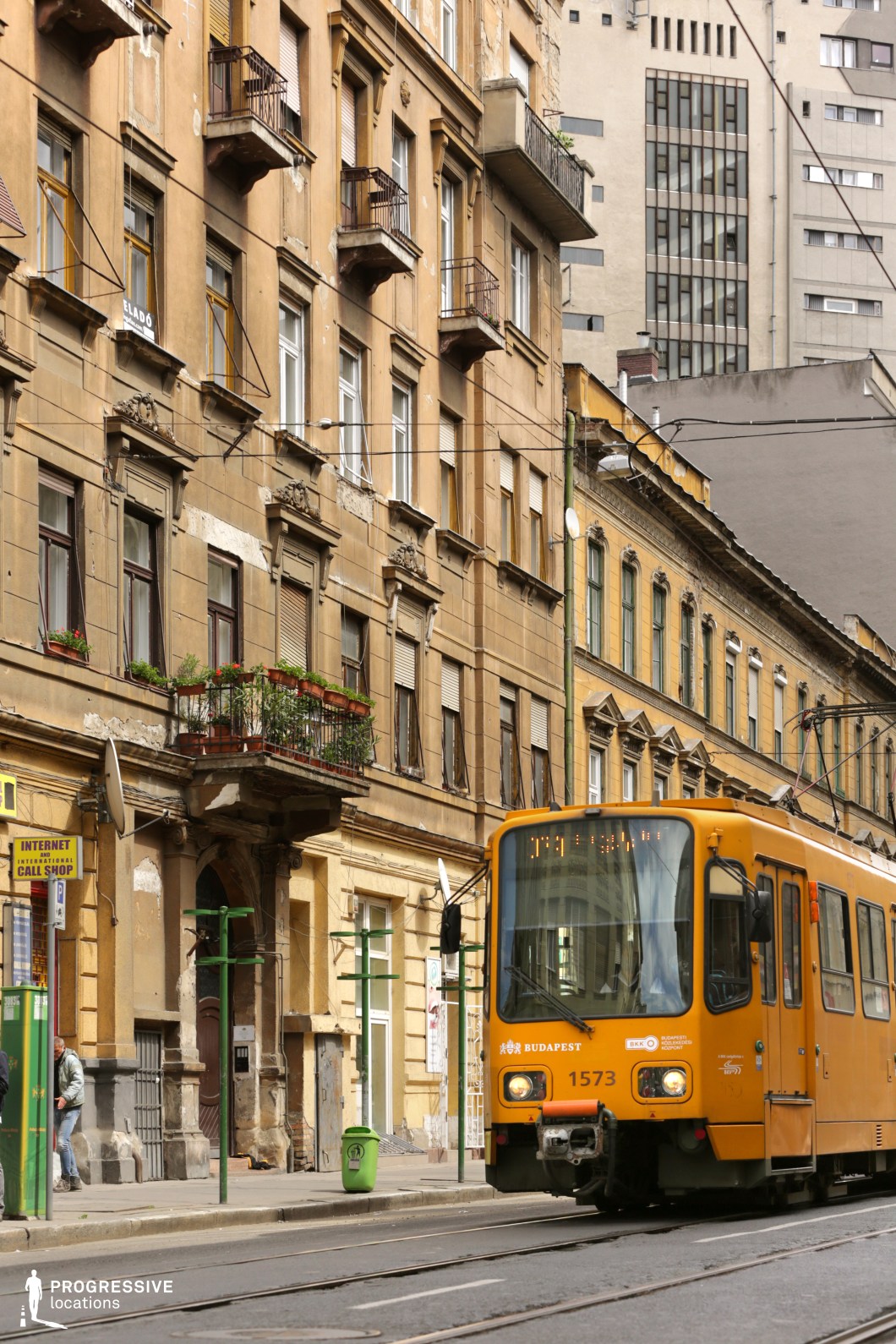 This image captures a classic Eastern European urban setting, with a yellow tram as the central element, running along the tram tracks that integrate seamlessly into the city street. The vibrant yellow of the tram creates a strong visual contrast with the aged and weathered facades of the surrounding buildings.

The buildings along the street exhibit ornate yet decayed classical architecture, with crumbling plaster, faded paint, and small, wrought-iron balconies filled with potted plants, adding a touch of local character. This combination conveys a sense of a city steeped in history and everyday life, providing an authentic urban texture.

In the background, modern buildings with sharp, minimalist designs break the historical continuity, adding another layer of contrast between old and new. This juxtaposition highlights the coexistence of architectural styles typical of many Eastern European cities.

The tram stop infrastructure, featuring simple green posts and signs, reflects the city's practical and utilitarian approach to public transportation. The street is narrow yet busy, making it suitable for sequences involving public transit, local movement, or urban commutes.

This location offers a distinctly lived-in, gritty atmosphere, ideal for productions requiring a realistic urban backdrop. The tram lines and overhead cables emphasize the integration of transportation into the urban fabric, adding elements of motion and direction to the scene.