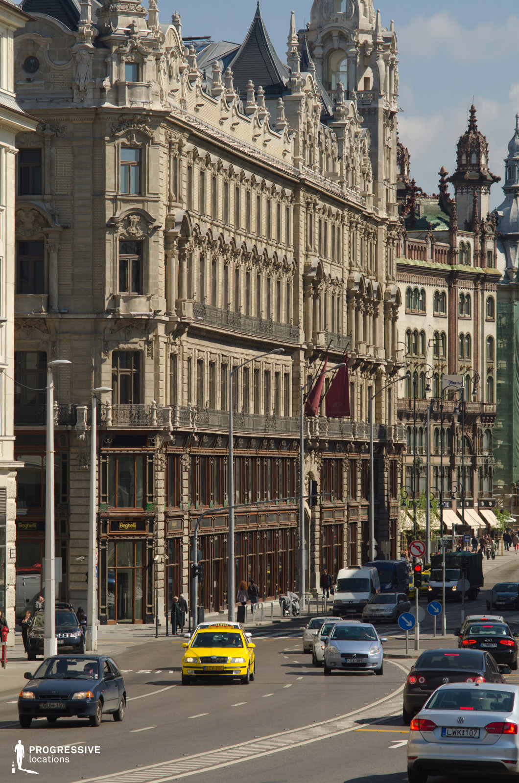 This wide city boulevard is flanked by majestic, five-story historic buildings featuring intricate architectural details. The facades are richly decorated with ornate stone carvings, arched windows, and decorative wrought-iron balconies, showcasing a grand European architectural style. The lower floors incorporate elegant wooden storefronts with large glass panels, adding a touch of modern functionality to the classical design. The street itself is wide and well-structured, accommodating multiple lanes of traffic, with a prominent yellow taxi providing a vibrant focal point. The scene reflects the sophistication of a bustling urban center, ideal for settings requiring a blend of historic grandeur and contemporary city life.