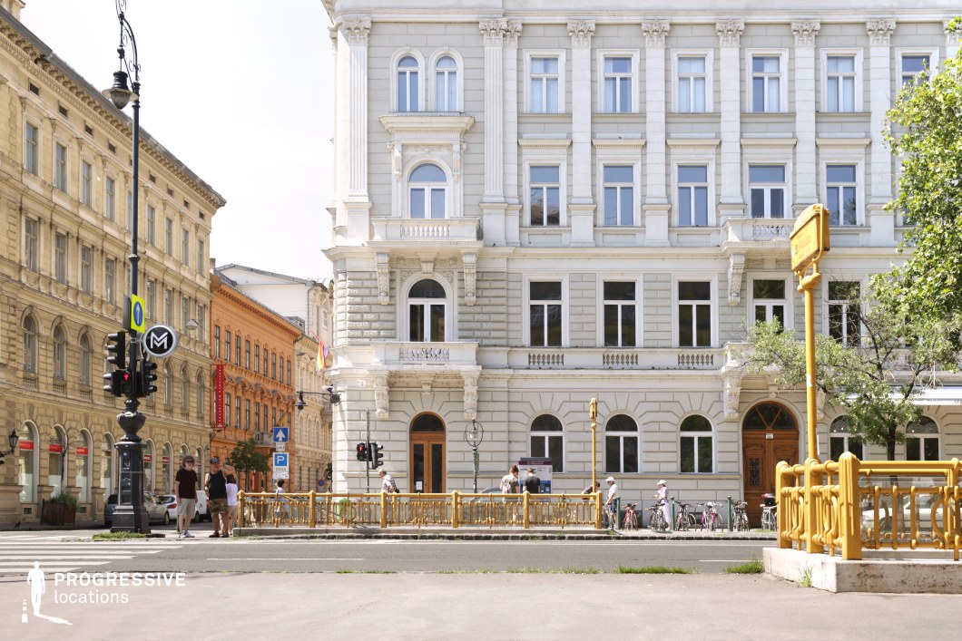 This bustling city intersection is framed by classic late-19th-century European architecture, with buildings in a neo-Renaissance style. Their pale stone facades are richly adorned with carved details, emphasizing symmetry and elegance. The standout feature is a wrought-iron railing enclosing a metro entrance, painted in a vibrant yellow—a distinctive touch that echoes the Art Nouveau tradition of integrating functional elements into decorative designs. The interplay of warm beige, soft greys, and the bold yellow creates a lively yet cohesive visual palette. The urban layout and architectural details evoke a strong resemblance to cities like Vienna or Paris, providing a dynamic yet charming backdrop for scenes of daily city life.