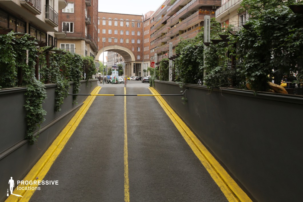 The image captures a narrow underpass leading to a visually striking archway framed by brick façades. The walls of the underpass are lined with lush greenery climbing up along planters, creating a sharp contrast between the industrial materials and the natural foliage. The yellow lane markings on the road guide the viewer's eye through the composition, reinforcing the perspective and depth. In the distance, the archway forms a strong focal point, framing a view of the street beyond. The surrounding buildings showcase a mix of brick and contemporary urban design elements, with balconies and terraces adding texture and layers to the scene. This setting is ideal for dynamic compositions, combining industrial and natural elements. The underpass and greenery offer an excellent backdrop for close-up photography, while the archway and surrounding urban environment provide opportunities for wide shots that emphasize perspective and symmetry.