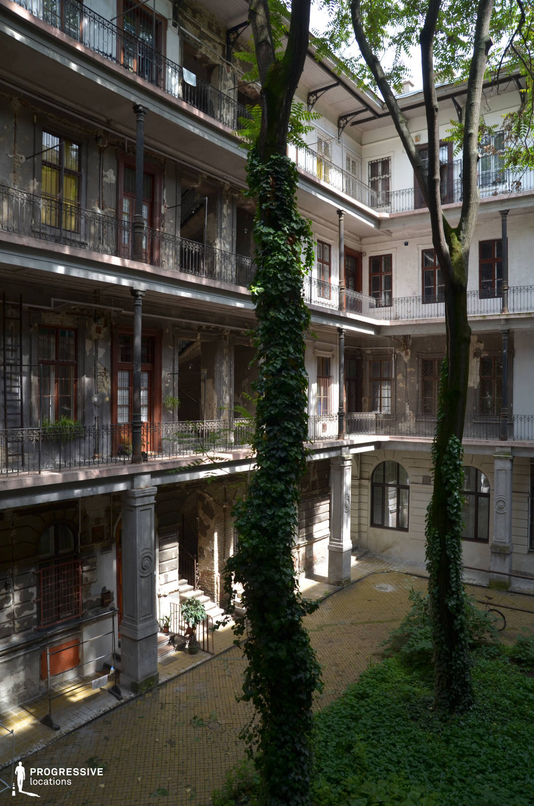 A picturesque yet weathered inner courtyard of a historic residential building, featuring multiple levels of wrought iron balconies supported by ornate columns. The aged facades display peeling paint and exposed brickwork, evoking a sense of old-world charm. Lush greenery, including ivy-clad trees and creeping vines, softens the architecture and adds a serene, slightly mysterious atmosphere. The cobblestone ground and arched entryways further enhance the location’s period aesthetic, making it ideal for period dramas, mystery narratives, or settings requiring an authentic European courtyard ambiance.
