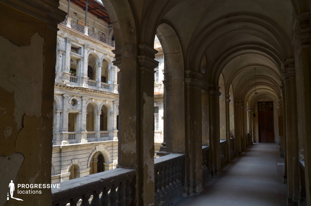 A grand, historic arcade overlooking a European-style courtyard with neoclassical influences. The long colonnade features a series of repetitive arches supported by robust columns, their surfaces showing aged patina and peeling plaster, adding to the location’s vintage charm. The balustrade offers a glimpse of the adjacent building, adorned with intricate stone arches, classical detailing, and wrought-iron balconies. Natural light filters through the openings, casting dramatic shadows that enhance the depth and texture of the space. This location is ideal for period dramas, architectural photography, or any production seeking an authentic, old-world European aesthetic.