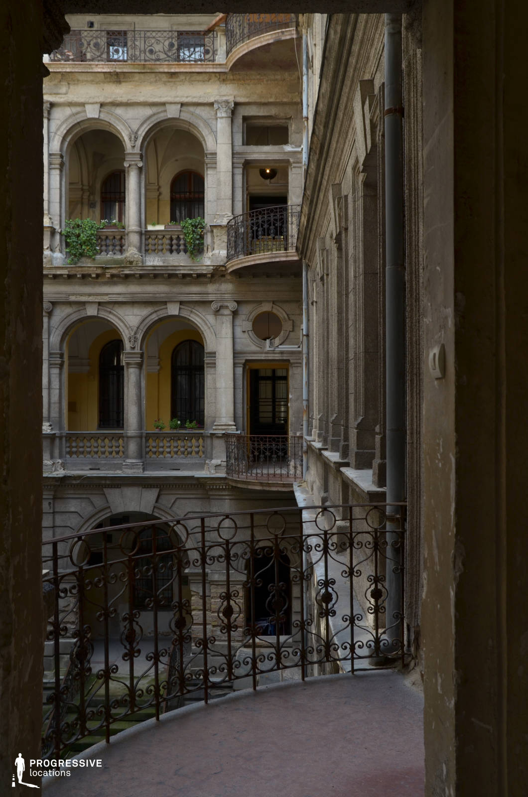 This magnificent historic courtyard features a layered architectural design with grand stone arches, classical columns, and decorative wrought iron railings. The aged stone façade, softened by creeping greenery and warm yellow accents, adds a timeless, slightly decayed elegance. The interplay of light and shadow enhances the depth of the space, emphasizing the intricate detailing on the balustrades and window moldings. The perspective from the curved balcony creates a dramatic framing effect, making this location ideal for filming period dramas, aristocratic intrigue, or mysterious noir thrillers. The rich textures and architectural grandeur evoke a sense of history, secrecy, and sophistication.