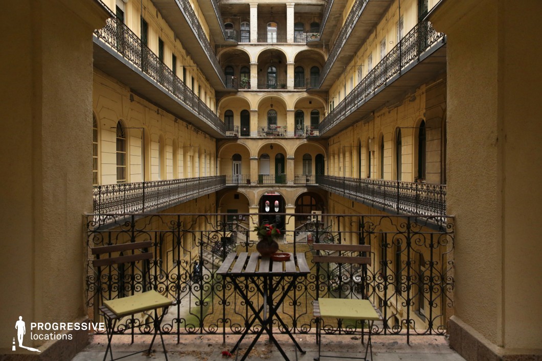 This grand, multi-story courtyard is enclosed by warm yellow-toned walls adorned with repeating arched windows and doorways, creating a sense of symmetry and classical elegance. Wrought-iron railings line the walkways on each level, adding an intricate decorative element. The central perspective emphasizes depth and balance, leading the eye toward the distant entrance framed by archways. The weathered facade and vintage architectural details evoke an old European atmosphere, making this location ideal for historical dramas, period films, or romantic narratives. The layered balconies and central void allow for dynamic cinematography, capturing movement and interactions from multiple vantage points.