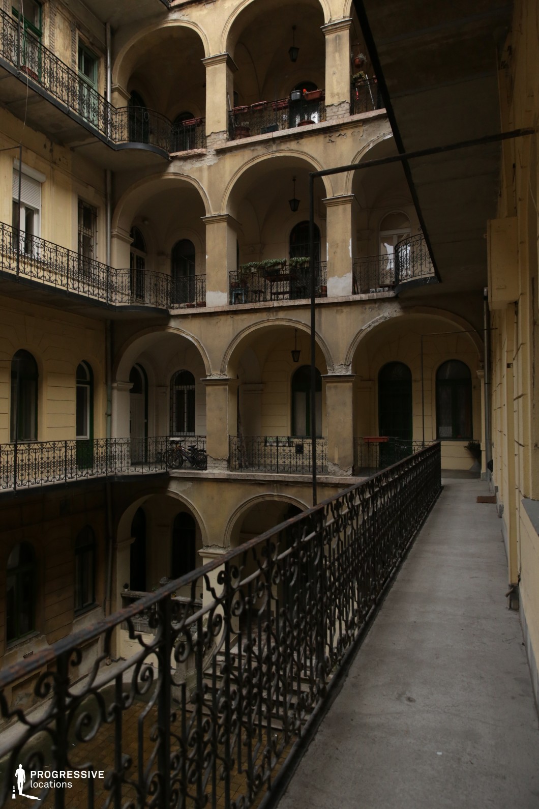 A multi-story courtyard framed by grand arches and wrought iron railings, evoking an old European charm. The aged yellow walls and weathered surfaces add a sense of history, while the open galleries provide depth and repetition, creating a striking visual rhythm. The space is rich in textures and architectural details, making it ideal for period pieces, noir-style films, or atmospheric dramas. The combination of intricate metalwork and classical arches allows for diverse lighting effects, from moody shadows to warm golden-hour illumination.