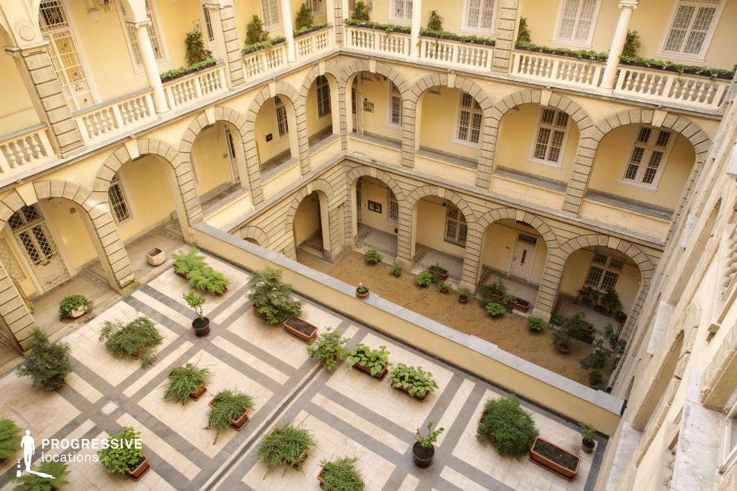 This elegant multi-story courtyard is framed by a symmetrical arrangement of neoclassical arches, textured stone accents, and pale yellow walls that evoke a refined, historical charm. Decorative balustrades and window grilles add an extra layer of sophistication, while the tiled ground level features an organized layout of lush potted greenery, creating a contrast between architectural formality and natural elements. The courtyard’s open-air structure and depth make it an ideal setting for period dramas, aristocratic intrigue, or romance films, offering a cinematic balance of grandeur and intimacy.