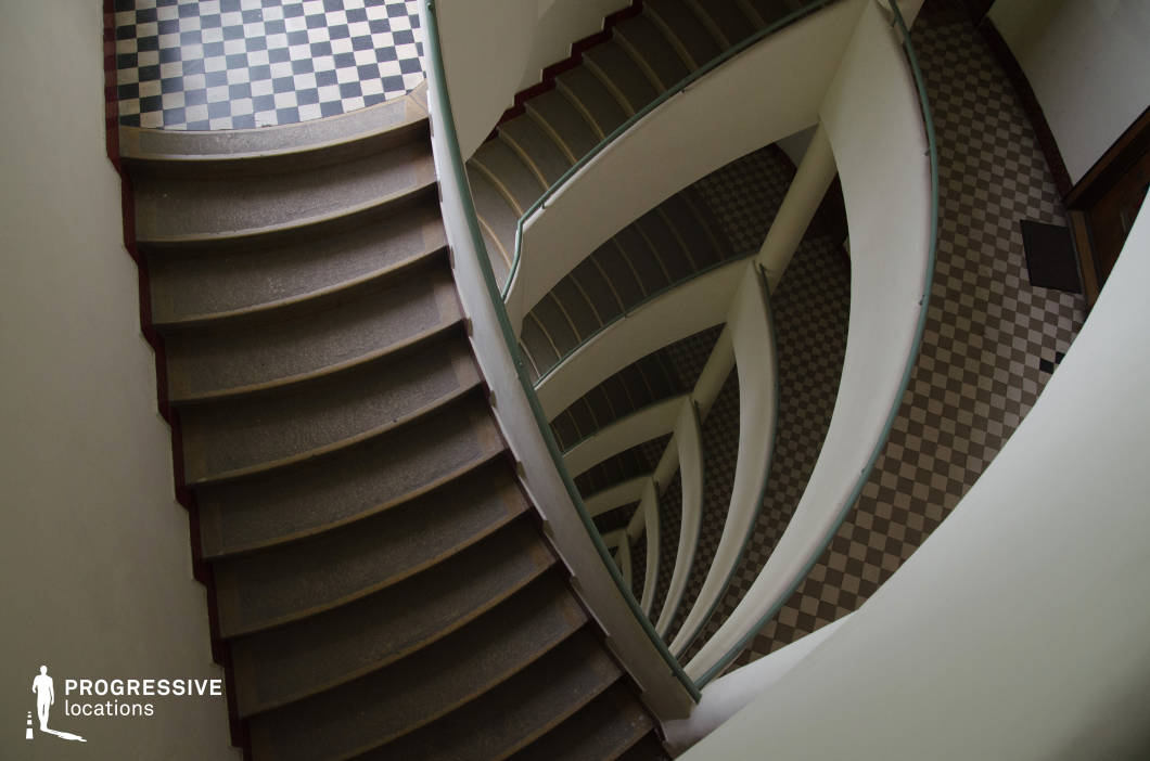 This dramatic spiral staircase features smooth, curved lines and a distinct Art Deco aesthetic. The sweeping railings, softened by pale surfaces, contrast with the bold black-and-white checkerboard flooring at both the top and bottom levels, reinforcing a sense of vintage elegance. The stairwell’s layered, repetitive structure creates a mesmerizing visual effect, enhanced by the interplay of light and shadow. The architectural fluidity and timeless design make this location ideal for psychological thrillers, film noir, or avant-garde cinematography, where dynamic camera movements can emphasize the staircase’s hypnotic depth and cinematic appeal.
