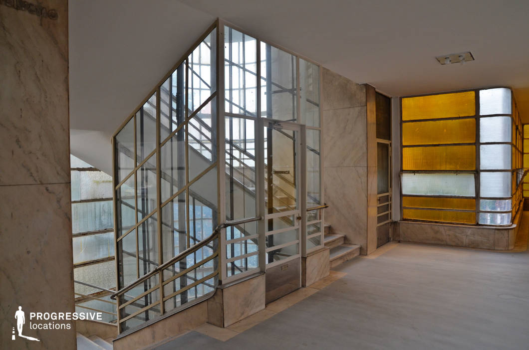 This striking Bauhaus-style stairwell features an abundance of geometric glass panels framed by thin metal railings, creating a structured yet elegant composition. The combination of frosted and amber-tinted windows allows diffused natural light to filter through, adding warmth to the cool marble surfaces. The glass-enclosed staircase, with its minimalist metal handrails and clean lines, reflects an early modernist aesthetic. This location is ideal for filming stylish period dramas, espionage thrillers, or retro-futuristic narratives, offering a sleek, sophisticated setting that blends functionality with artistic design.