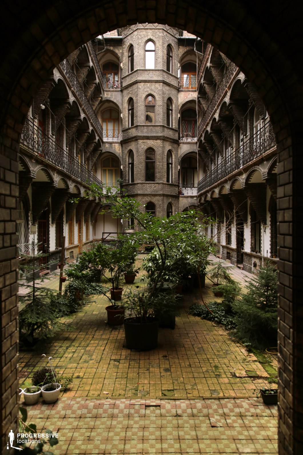 A grand, enclosed courtyard framed by an arched brick entryway leads into a striking multi-story building with intricate wrought-iron balconies and arched walkways. The aged brick facade, adorned with symmetrically placed windows and decorative ironwork, evokes a historic European architectural style. The courtyard is paved with worn, moss-tinged tiles and is lush with potted plants, adding a natural contrast to the rigid geometry of the architecture. The vertical symmetry and depth of the scene create a visually immersive setting, making it ideal for period dramas, mystery films, or noir-inspired cinematography. The secluded atmosphere and interplay of light and shadow enhance its potential for intimate or suspenseful scenes.