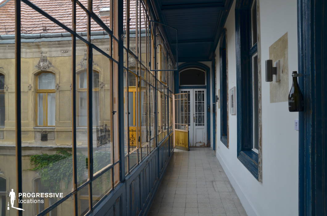 This image features an enclosed loggia in a historic residential building, with large glass windows framed by aged blue wooden structures. The worn paint and slight weathering on the surfaces add a sense of character and history. The narrow tiled corridor is bordered by white walls with decorative blue moldings, leading to an arched doorway with intricate glass-paneled doors. The windows reflect the opposite façade, revealing ornate architectural details, warm yellow window frames, and ivy creeping along the building, reinforcing the old-world charm. This visually layered space is ideal for filming period pieces, romantic dramas, or introspective character-driven narratives, offering a mix of nostalgia and urban elegance.