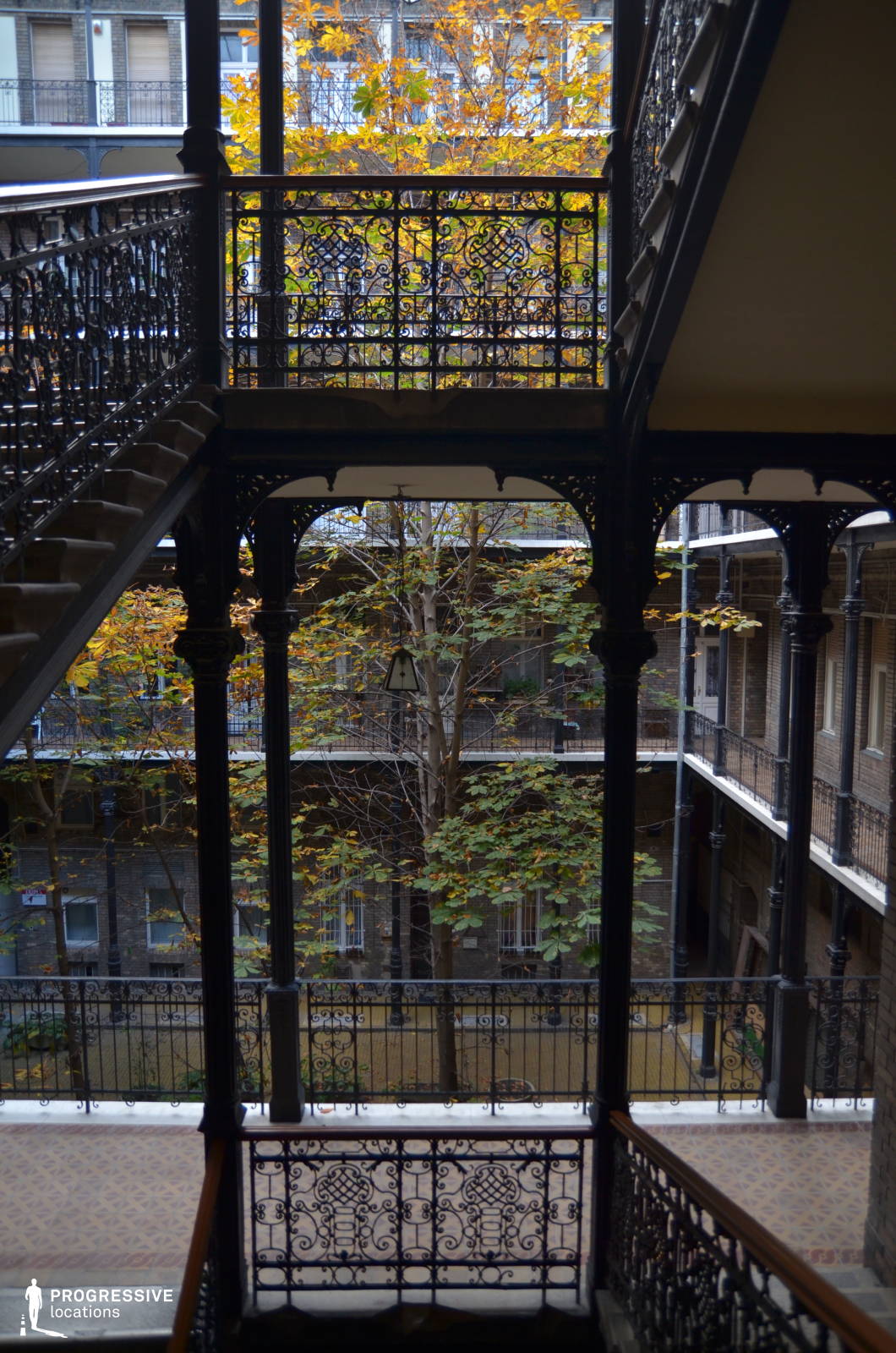 This picturesque courtyard features an intricate cast-iron staircase and balcony railings, exuding a classic European charm. The symmetrical, open-air structure allows natural light to filter through, highlighting the rich textures of the aged brick walls and the delicate ironwork details. The layered walkways and central tree create a visually dynamic setting with depth and character. This location is ideal for filming period dramas, romantic narratives, or noir-inspired thrillers, offering an evocative blend of old-world elegance and atmospheric intimacy.