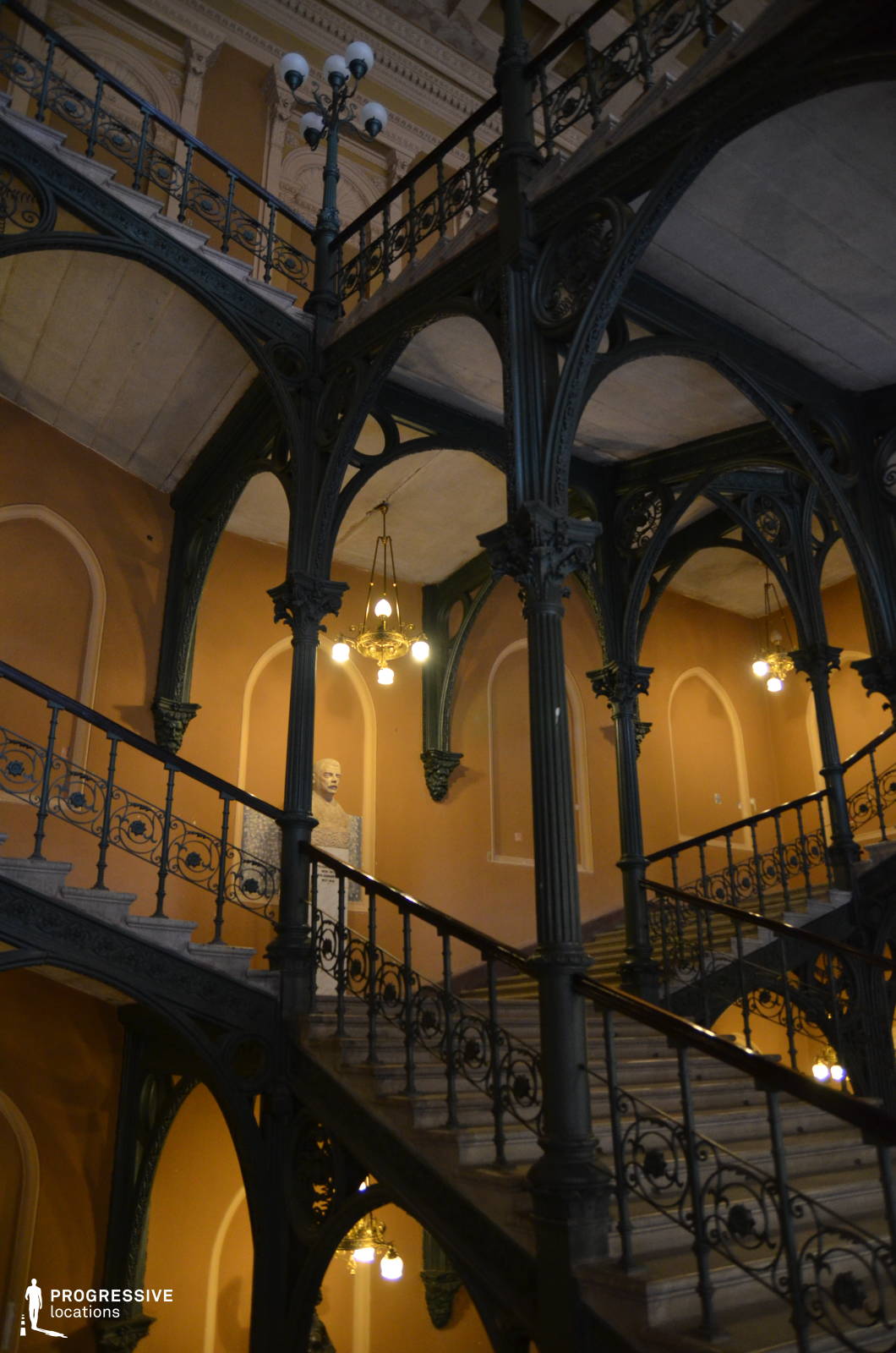 This striking multi-level staircase is framed by intricate cast-iron columns and railings, creating a dramatic interplay of structure and ornamentation. The deep green metal framework, adorned with elaborate archways and decorative flourishes, contrasts beautifully with the warm ochre walls and arched recesses. Classic chandeliers cast a soft glow over the stone steps, emphasizing the building’s historical grandeur. This visually rich setting is ideal for period dramas, gothic thrillers, or mystery films, offering a cinematic blend of elegance, architectural mastery, and a timeless, slightly mysterious atmosphere.