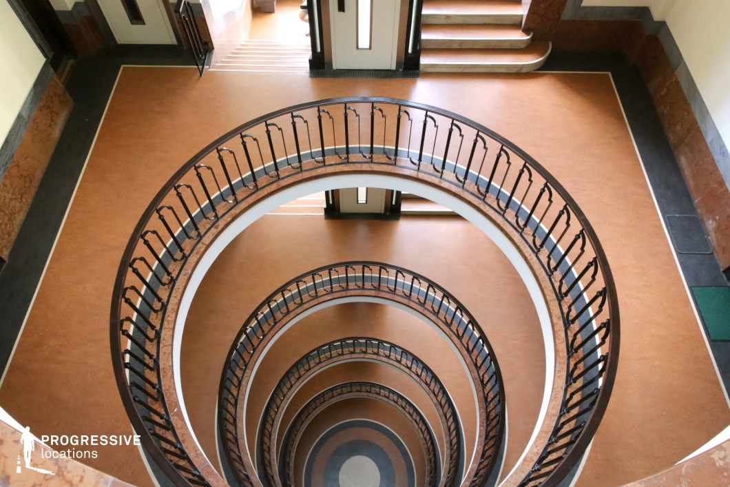 This grand circular staircase spirals downward in a mesmerizing pattern, framed by gracefully curved metal railings with subtle Art Deco influences. The warm-toned flooring, accented with marble details, enhances the sophisticated and timeless aesthetic of the space. The layered symmetry and smooth transitions between floors create a visually captivating depth, while the polished surfaces reflect soft natural light. The combination of refined materials and architectural precision makes this location ideal for period dramas, high-society thrillers, or stylish noir films, offering a striking and immersive backdrop for cinematic storytelling.