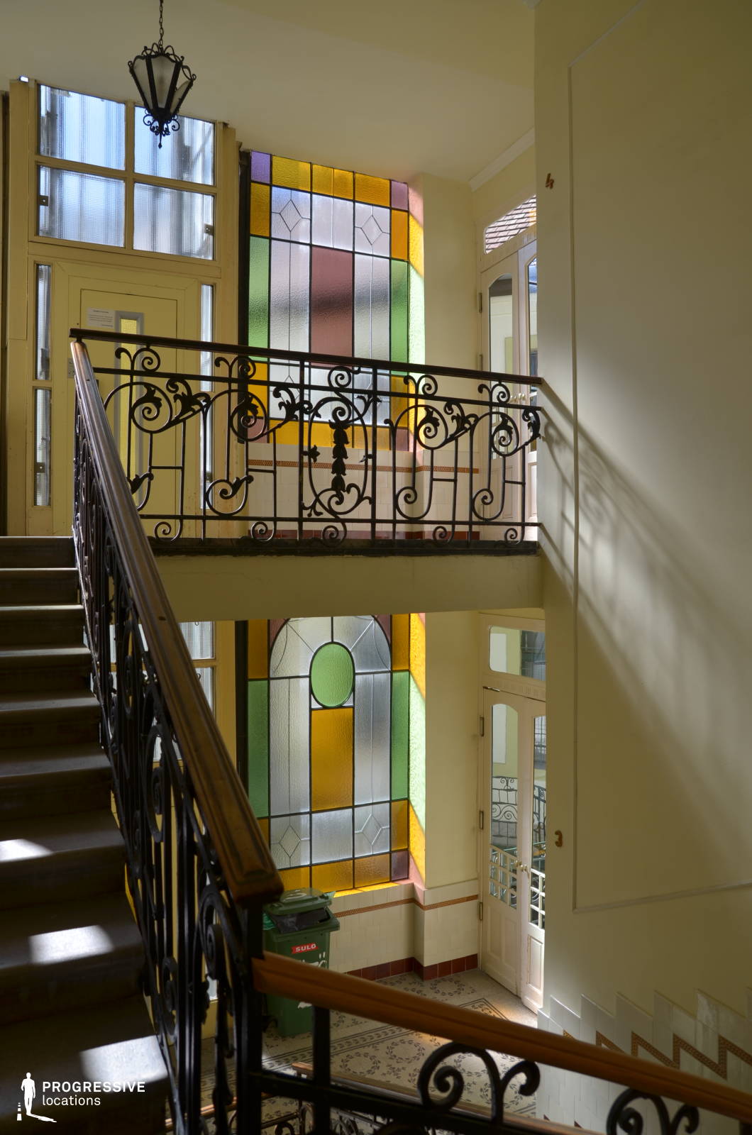 This beautifully lit stairwell is adorned with intricate wrought-iron railings and striking stained-glass windows that cast warm, multicolored light onto the walls and floor. The soft cream-colored walls and delicate tilework contribute to the refined atmosphere, while the natural light filtering through the glass enhances the sense of openness. The combination of elegant curves and geometric patterns gives the space a timeless charm. This location is ideal for romantic dramas, period pieces, or artistic films, offering a luminous and inviting setting that balances vintage sophistication with a dreamy aesthetic.