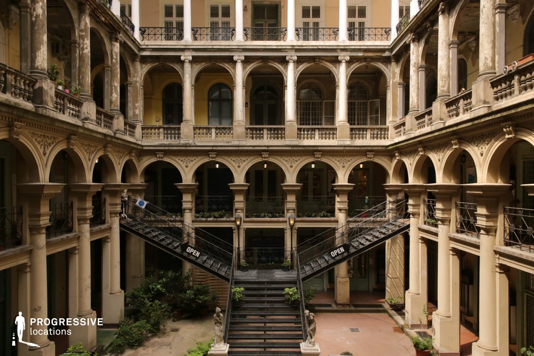 This impressive multi-story courtyard features a rich blend of classical and industrial elements, with grand arched colonnades, weathered stone balustrades, and an imposing symmetrical iron staircase. The faded elegance of the architecture, with its intricate decorative carvings and aging surfaces, evokes a sense of historical grandeur. Lush greenery softens the formal structure, while the dramatic dual staircases leading to the upper levels create a visually striking focal point. This location is ideal for period films, historical dramas, or gothic mysteries, offering an atmospheric and immersive backdrop that enhances themes of intrigue and timeless sophistication.