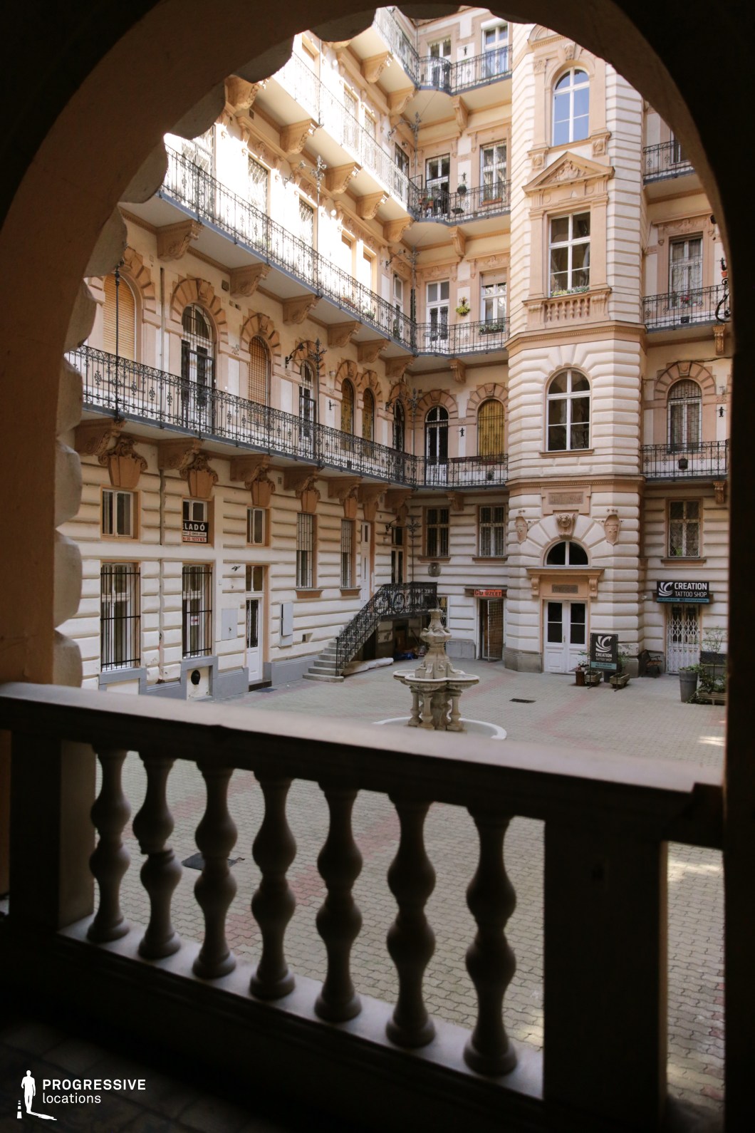 This opulent courtyard, framed by an arched balustrade in the foreground, showcases a richly decorated multi-story facade with intricate stucco moldings, sculpted window surrounds, and elegant wrought-iron balconies. The warm-toned exterior features classical detailing, including pilasters and ornamental reliefs, while a central fountain adds a refined focal point to the open space. The combination of decorative staircases and tiered balconies creates a dramatic sense of depth and sophistication. This location is ideal for period dramas, aristocratic intrigue, or romantic films, offering a setting of timeless elegance and European grandeur.