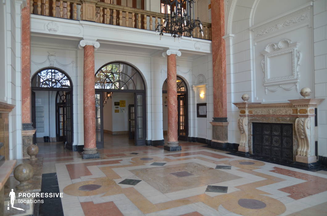 A luxurious entrance hall featuring a blend of classical and baroque influences, with white sculpted walls, decorative moldings, and striking red marble columns. The intricately designed floor showcases geometric marble inlays, adding a sophisticated touch. A grand fireplace with elaborate carvings anchors the space, while an imposing wrought-iron chandelier hangs above, enhancing the atmosphere. Arched doorways with black metal frames provide a contrast to the predominantly white interior, allowing for elegant depth. This opulent setting is ideal for period dramas, high-end commercial shoots, or historical productions requiring a lavish backdrop.
