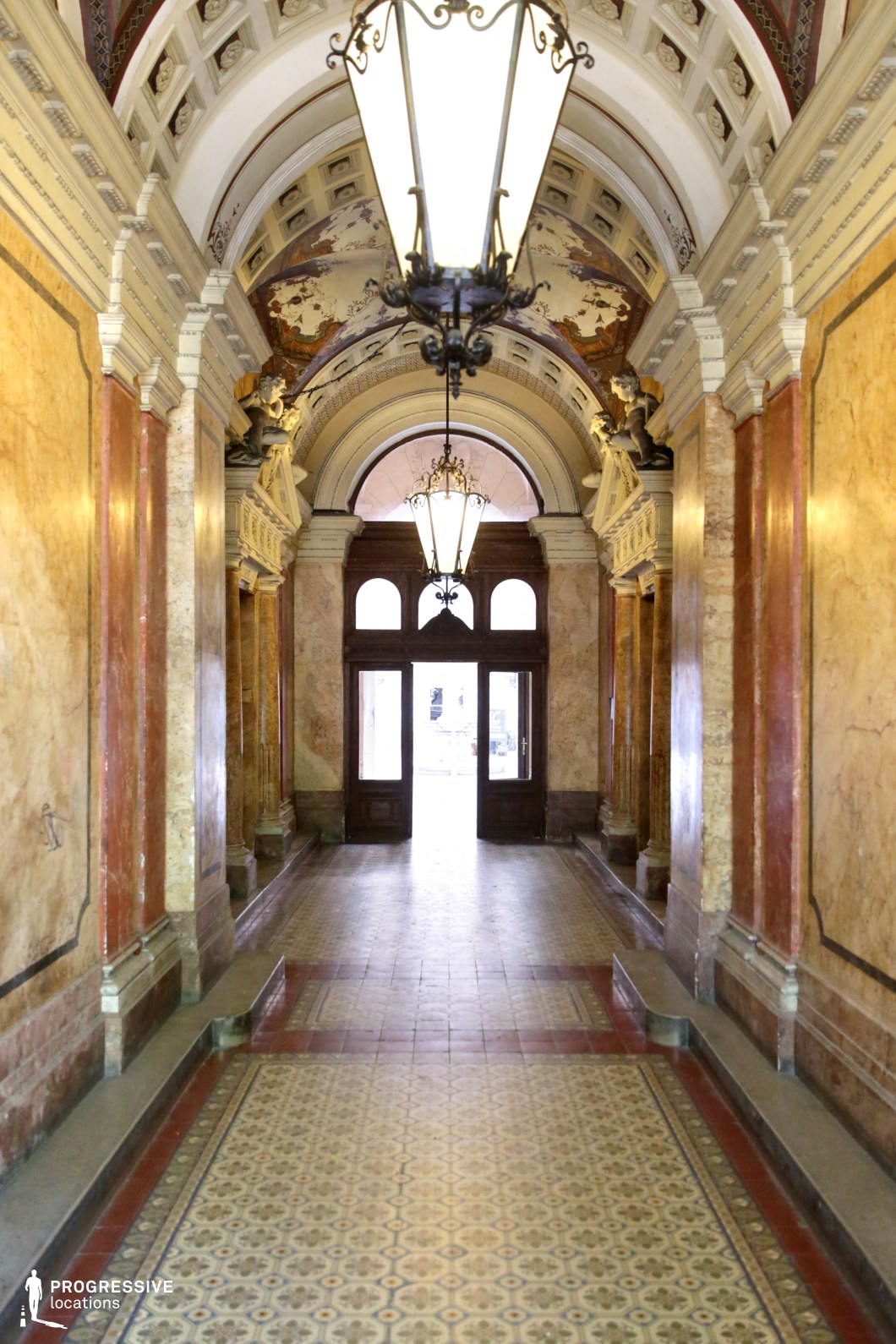 This grand neoclassical entrance hall is adorned with richly veined marble walls, intricate vaulted ceilings, and elegant sculptural reliefs. The warm tones of the stone contrast beautifully with the patterned tile flooring, leading the eye toward a grand arched doorway framed by dark wooden panels. Large wrought-iron chandeliers hang from the ceiling, casting a soft glow that enhances the opulent yet aged atmosphere. The finely detailed architectural elements make this space ideal for period dramas, historical intrigue, or gothic thrillers, offering a sophisticated and immersive setting that evokes old-world elegance and mystery.