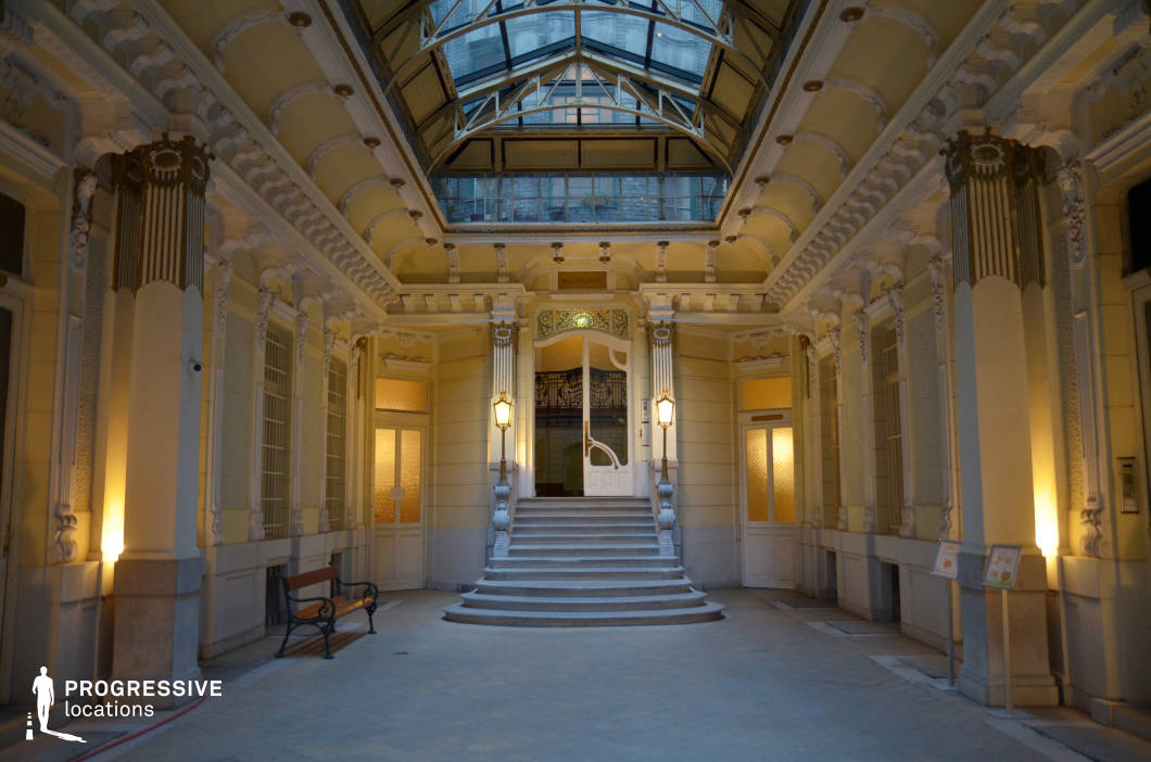 This grand Art Nouveau entrance hall exudes sophistication, featuring ornate decorative moldings, fluted pilasters, and intricate sculptural embellishments. The pale yellow walls and curved architectural elements create a soft, welcoming atmosphere, further enhanced by the warm glow of classical lanterns. A dramatic glass ceiling framed with wrought iron allows natural light to filter in, adding an airy elegance to the space. The gently curved staircase, leading to a decorative glass-paneled doorway, serves as a focal point. This visually striking location is ideal for period dramas, high-society intrigue, or romantic films, offering a refined and cinematic setting filled with timeless charm.