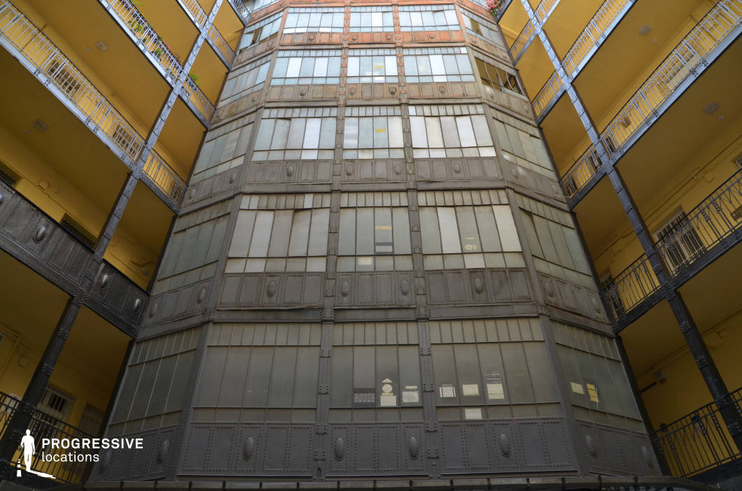 This imposing courtyard features an industrial-era iron framework, combining riveted metal panels with frosted glass to create a strikingly structured, symmetrical facade. The contrasting warm yellow walls and delicate wrought iron railings add a softer, almost theatrical feel to the otherwise rigid, factory-like architecture. The multi-level balconies frame the central iron-and-glass structure, emphasizing depth and perspective. This location is ideal for filming neo-noir thrillers, dystopian sci-fi, or historical dramas, offering a dramatic backdrop that blends industrial elegance with an air of mystery and urban grandeur.