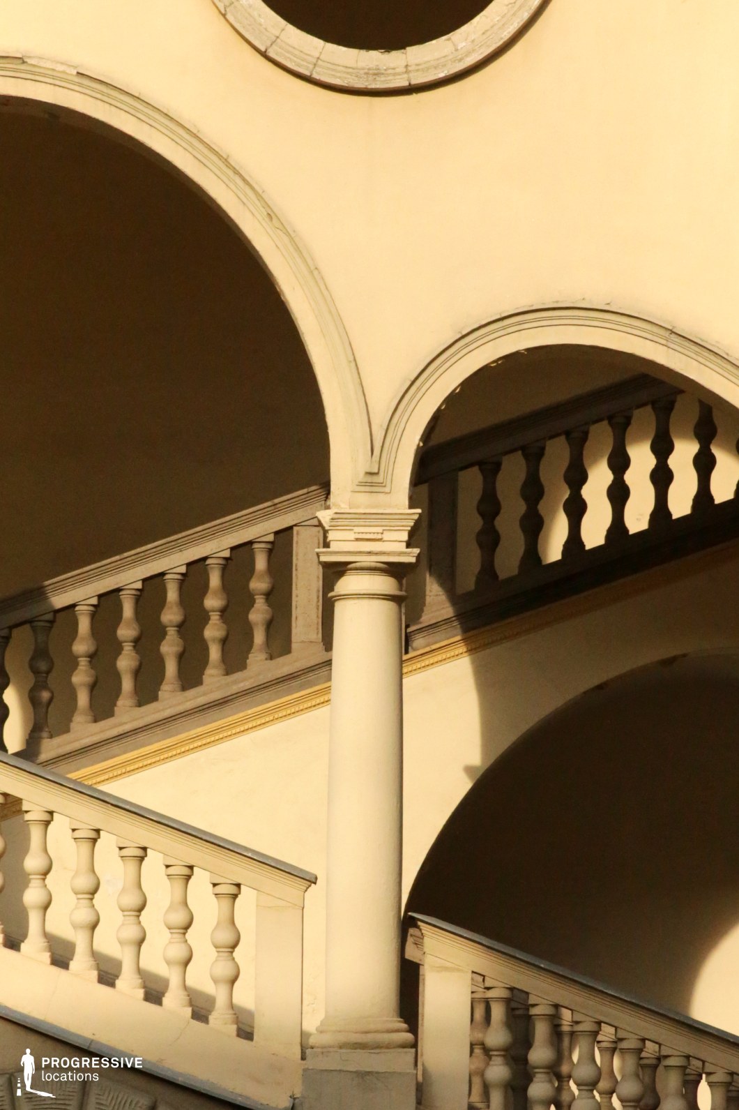 This neoclassical architectural composition features a series of gracefully layered arches, intricate balustrades, and a single column supporting the intersecting stairways. The warm-toned facade is highlighted by elegant detailing, including smooth curved moldings and sculpted balusters that add depth and refinement. The play of light and shadow enhances the three-dimensional quality of the space, creating a visually compelling contrast between structure and void. This location is well-suited for period dramas, historical narratives, and elegant mystery films, offering a timeless and sophisticated setting with a cinematic balance of geometry and classical grandeur.