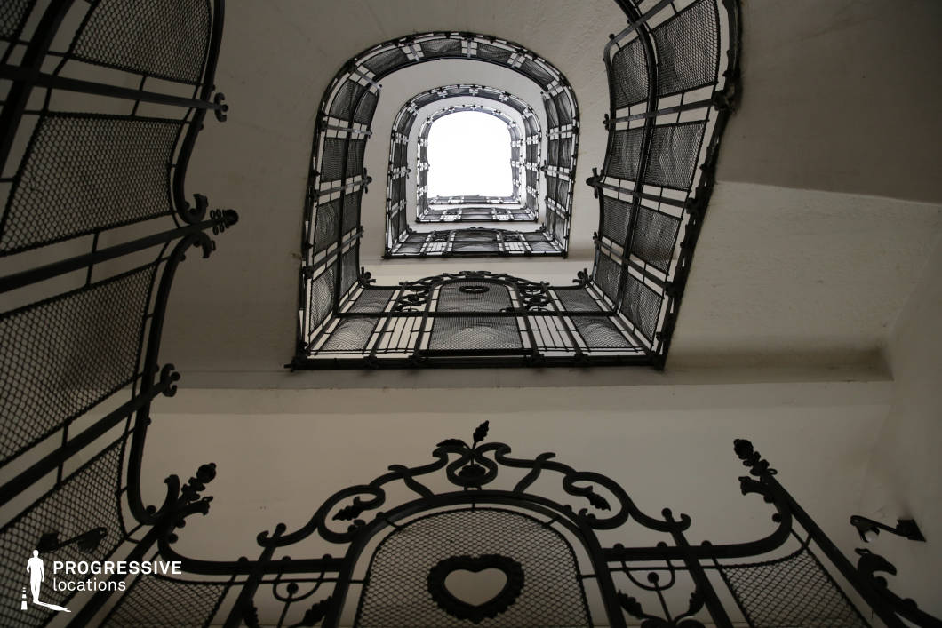 This striking stairwell features intricate wrought iron railings adorned with delicate scrollwork and geometric mesh panels, creating a visually rich interplay of light and shadow. The ascending levels form a dramatic perspective, framed by white walls that contrast sharply with the dark metal detailing. The repetition of arched openings adds depth and symmetry, reinforcing the stairwell’s architectural elegance. The heart-shaped motif in the foreground hints at an Art Nouveau influence. This location is ideal for filming period dramas, gothic thrillers, or historical mysteries, offering a refined yet slightly enigmatic setting that enhances atmosphere and visual storytelling.