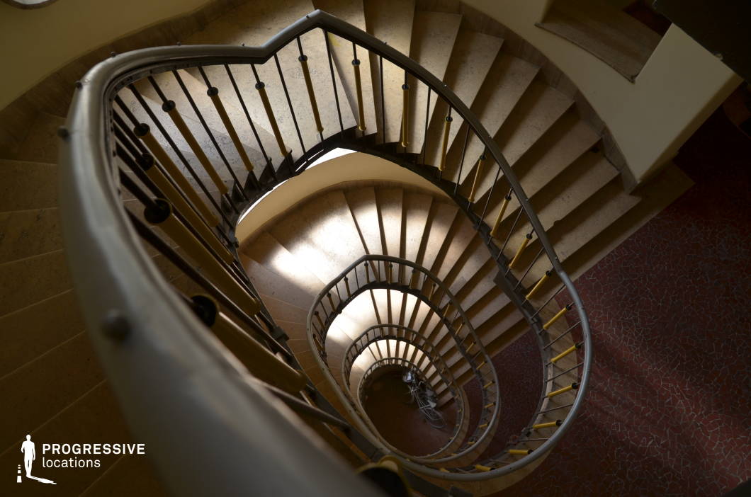 This image captures a striking spiral staircase with a smooth, curving metal railing and a series of evenly spaced vertical balusters, leading the eye downward in a hypnotic visual rhythm. The worn stone steps contrast with the sleek metal elements, while soft, directional light casts subtle shadows, enhancing the depth and curvature of the structure. The surrounding walls follow the organic flow of the staircase, reinforcing the architectural harmony. The patterned red-tiled floor at the base adds a textured contrast to the neutral tones above. This setting is ideal for filming psychological thrillers, noir-style mysteries, or dramatic sequences emphasizing tension, movement, and perspective.
