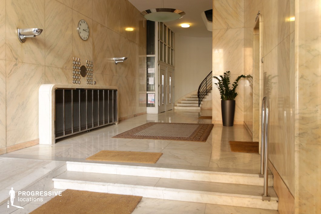This sophisticated lobby features a sleek mid-century modern aesthetic, with walls and floors clad in polished yellow-white marble that exudes luxury. The symmetrical design is accentuated by geometric patterns, including a striking circular button panel and metallic wall sconces. The floating staircase with a minimalist black railing curves upward, adding a sense of openness and fluidity. Soft lighting fixtures and decorative rugs complement the refined atmosphere. This location is ideal for filming stylish period dramas, espionage thrillers, or high-society narratives, offering a pristine yet subtly mysterious setting that enhances intrigue and elegance.