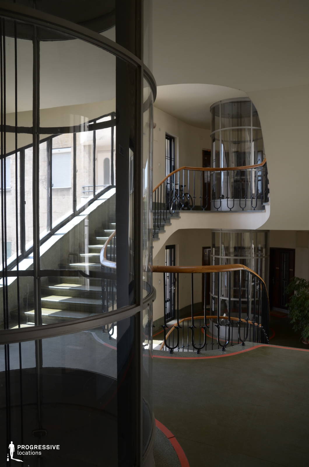 This modernist staircase wraps elegantly around a cylindrical glass elevator, creating a visually dynamic composition of curves, reflections, and open space. The smooth, flowing railings with warm wooden handrails contrast with the cool-toned flooring, which is subtly accented with red outlines. Natural light filters through the large windows, enhancing the architectural depth and highlighting the interplay of transparency and structure. The layered design and fluid movement of the staircases make this location ideal for filming stylish period pieces, retro-futuristic narratives, or psychological thrillers, offering a sophisticated and almost surreal aesthetic.