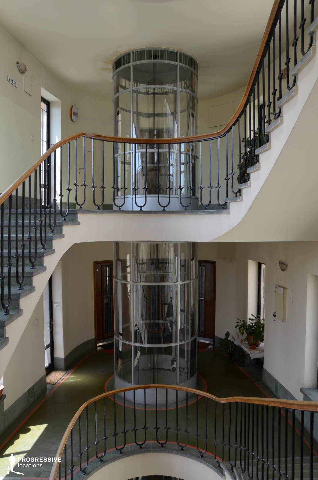 This striking interior showcases a central cylindrical glass elevator encased in a sleek metal frame, surrounded by a gracefully curving staircase with wrought iron railings and polished wooden handrails. The light-colored walls contrast with the deep green flooring, accentuated by subtle red outlines that add a mid-century modern touch. The symmetry of the space, combined with the transparency of the elevator, creates a dynamic visual effect, emphasizing openness and movement. This setting is ideal for filming stylish period dramas, retro-futuristic narratives, or suspenseful thrillers, offering a refined yet slightly mysterious ambiance reminiscent of classic European architecture.