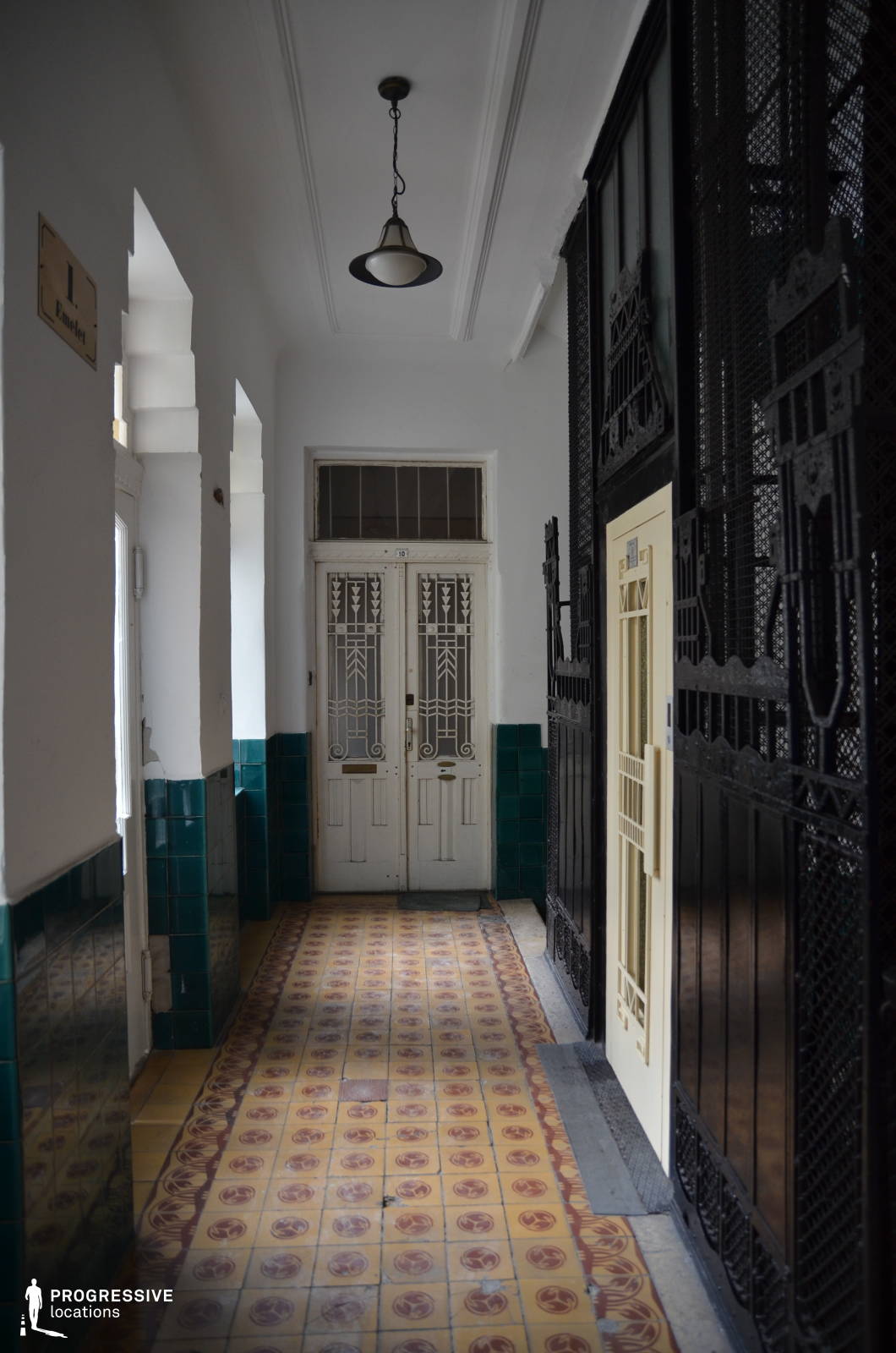 This narrow stairwell captures a distinct early 20th-century charm, featuring patterned ceramic floor tiles in warm tones, deep green glazed wall tiles, and intricate wrought iron elevator grilles. The vintage white double doors with decorative glass panels add an element of refinement, while the black metal framework of the elevator shaft enhances the contrast between elegance and industrial craftsmanship. The soft lighting and aged textures contribute to a nostalgic, cinematic atmosphere. This location is ideal for filming period dramas, noir thrillers, or mystery narratives, offering a richly detailed and immersive historical setting.